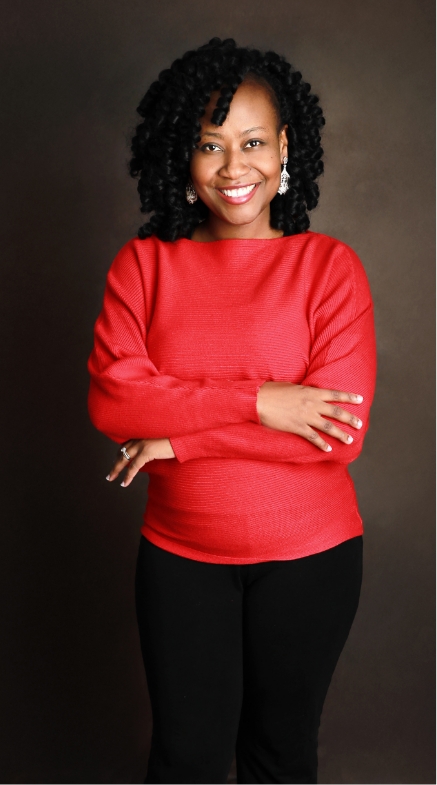 A woman with a bright smile wearing a red sweater and black pants, standing with her arms crossed against a brown background.