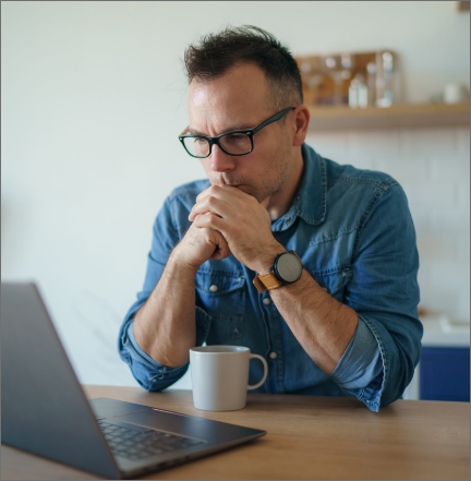 Man with glasses concentrating on laptop screen, with a cup of coffee at his workspace.