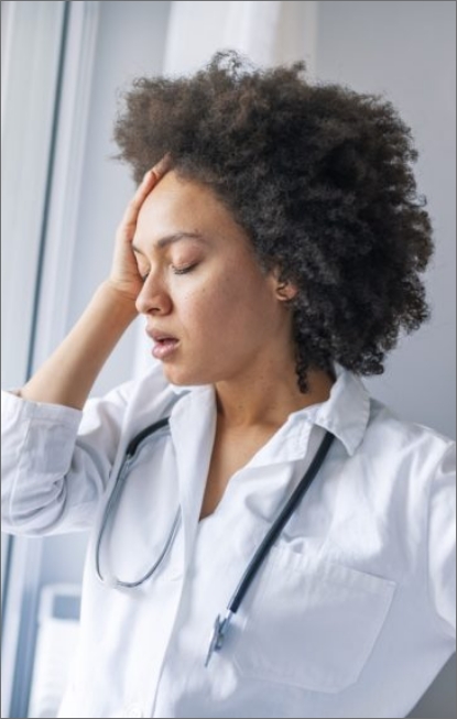 A healthcare professional appears fatigued or stressed, touching her forehead in a gesture of concern or exhaustion.