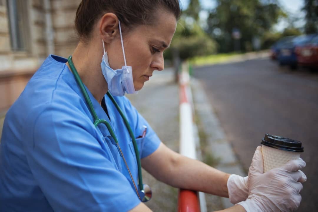 Physician experiencing burnout symptoms sitting outside hospital holding coffee after long shift